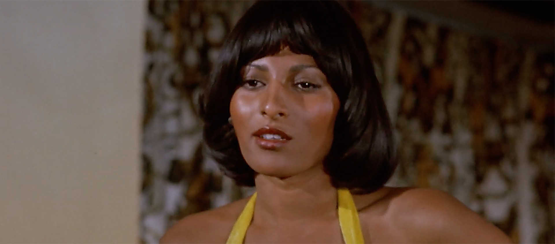 Pam Grier from the shoulders up. She has medium brown skin and a dark brown bob with bangs and wears a yellow halter top.