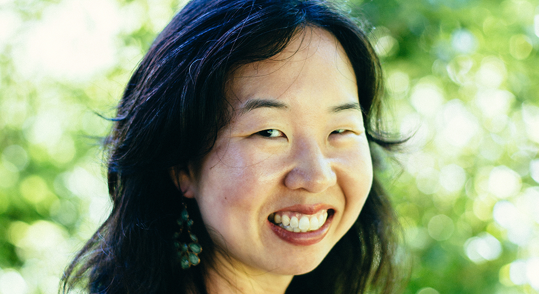 Close-up shot of Shelley Wong, who has wavy black hair and light skin, in front of a blurred background of greenery and light. 