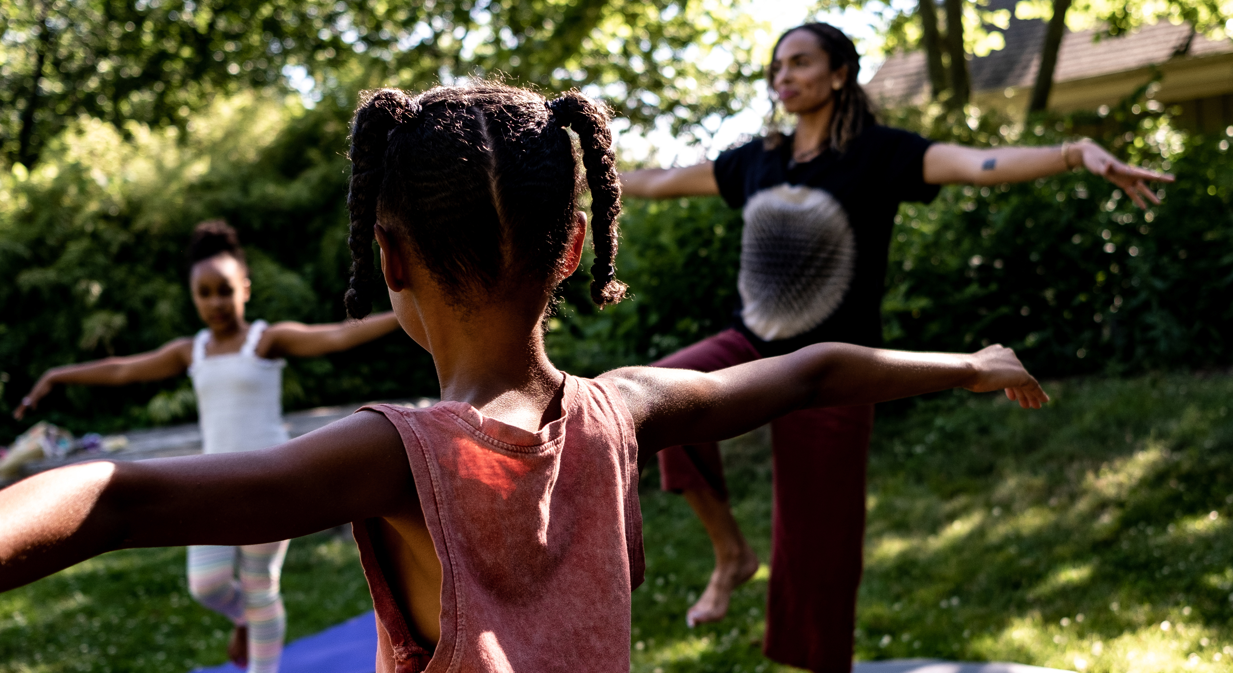 Two children and an instructor standing outside in grass and holding out their arms in yoga poses. There are trees and bushes in the background.