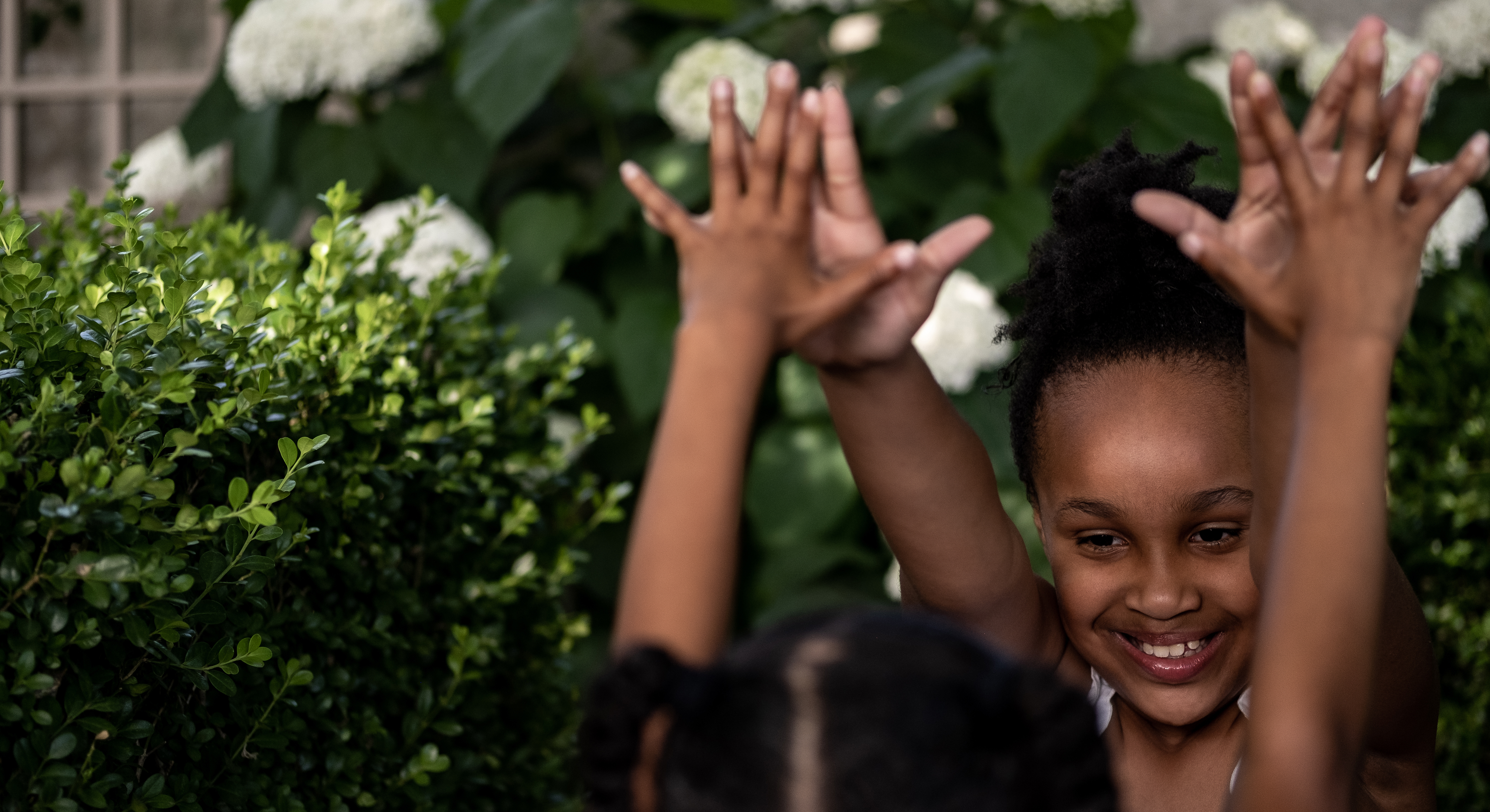 Two children with medium brown skin and dark hair; they are surrounded by greenery and facing each other with their arms raised and fingertips touching.