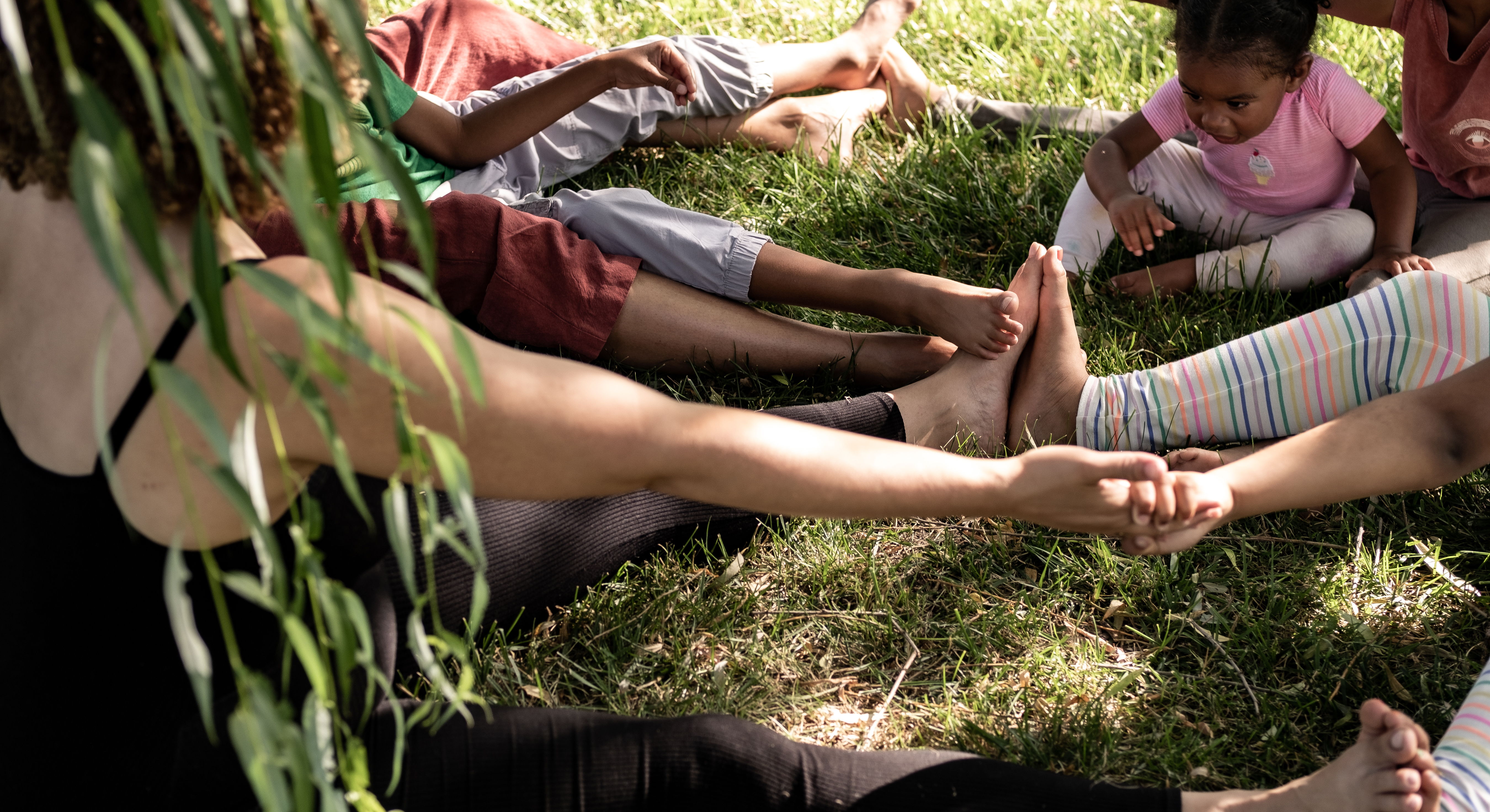Group of children and adults sitting in grass with legs and arms outstretched. Two people are holding hands.