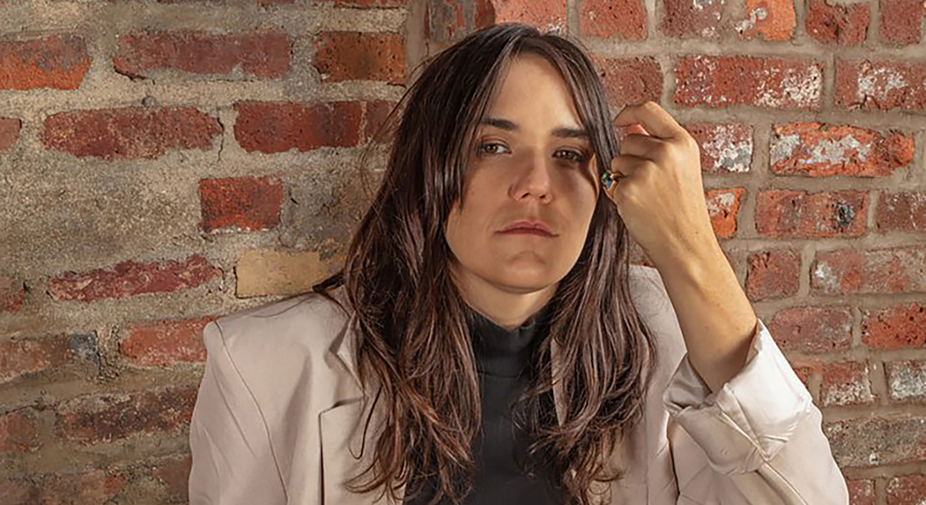 Artist in a beige suit sits on top of a chair in front of a brick wall