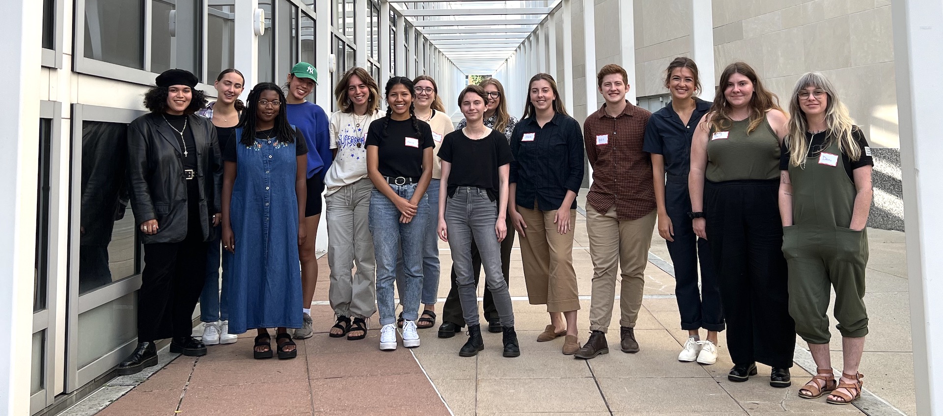 A lineup of 14 student interns working at the Wexner Center for the Arts in 2022-23. They're standing in a walkway between pillars of a white metal grid that's adjacent to the center building.
