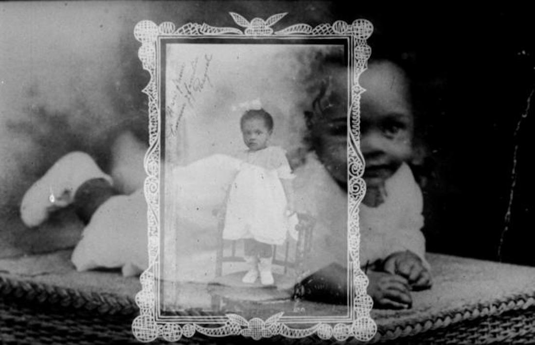 Still from the Sara Gómez film Crónica de mi familia. A black and white image of a Black baby in white dress and booties lies on its stomach on a wicker basket. A photographed portrait of a slightly older Black child in similar clothing, standing alone, is superimposed over the image of the baby.