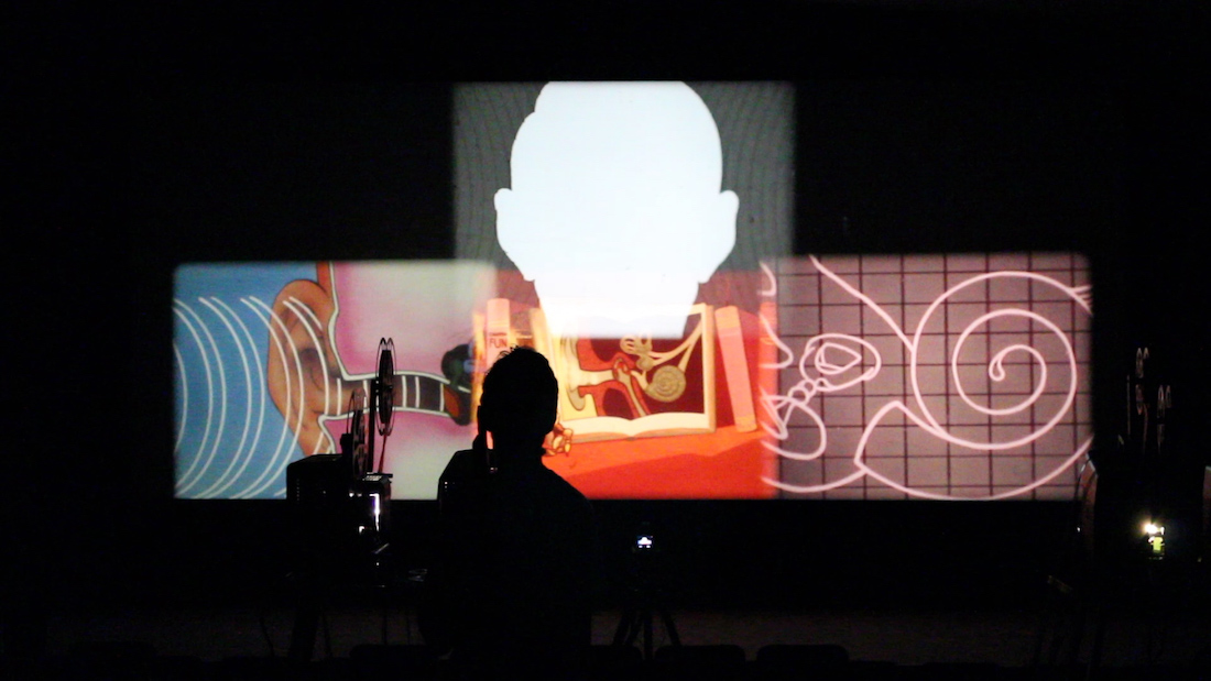 An image of an experimental film performance. A collage of four projected images that includes a human head in white silhouette and illustrations of the inner ear are seen on a wall in a dark room. The silhouette of a man operating a 16 millimeter projector is near the center.