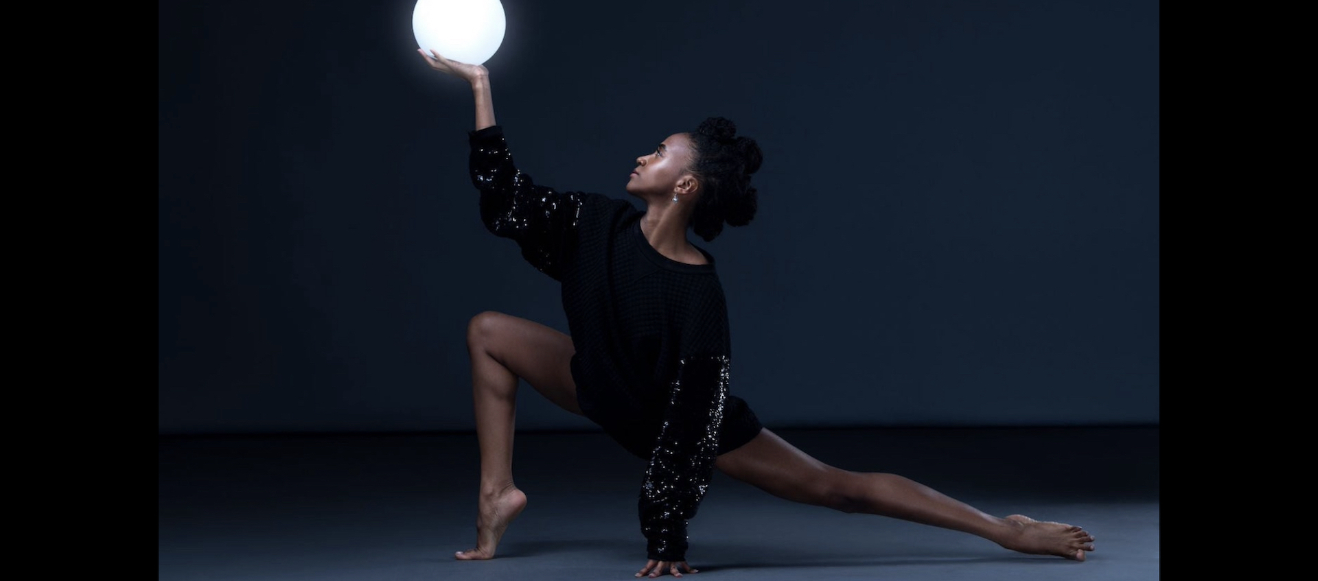 A female member of the Ailey II dance company is alone on a darkened stage, wearing a short black dress with long, sequined sleeves. She is lunging low to her right with her left hand on the floor. Her right hand is raised up in the air, holding an illuminated white orb.