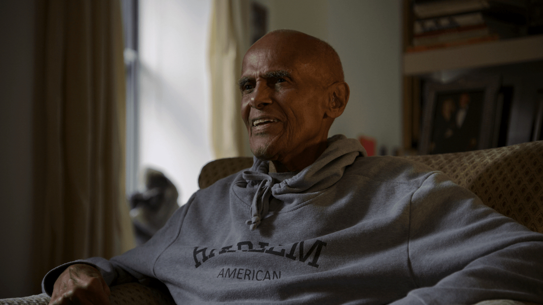 Still from the documentary Is It Black Enough for You? Actor Harry Belafonte sits in a living room wearing a gray sweatshirt. He looks to his right, smiling.