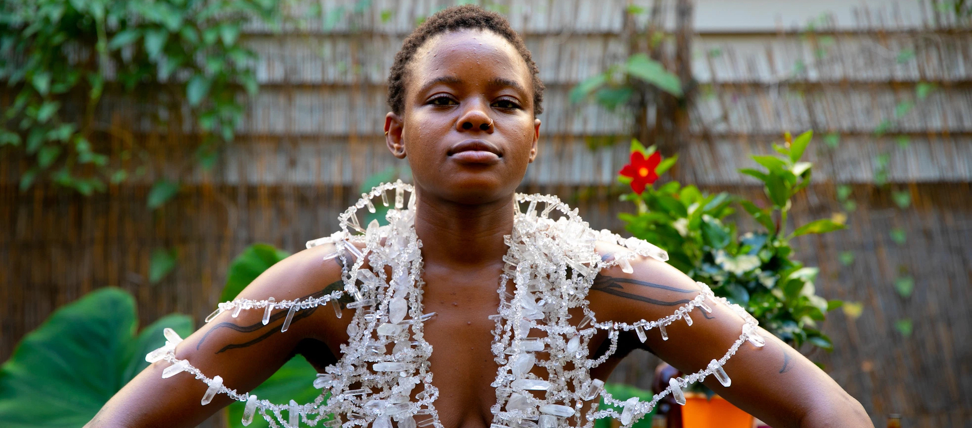 A young Black woman with short natural hair sits outdoors. There are plants and a cottage wall behind her. She is wearing only a large necklace of white stones, which covers her torso and drapes over her upper arms.