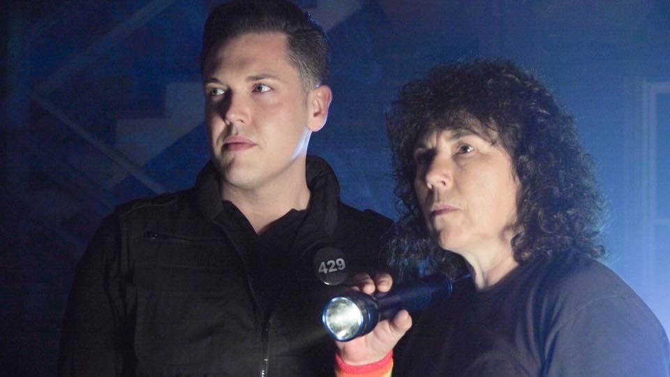 Queer Ghost Hunters Shane McClelland and Lori Gum. He is a white man with short dark hair. She is a white woman with shoulder-length curly hair. Both are wearing all black And staring into the distance to their left. She is holding a powered-up flashlight in that direction.