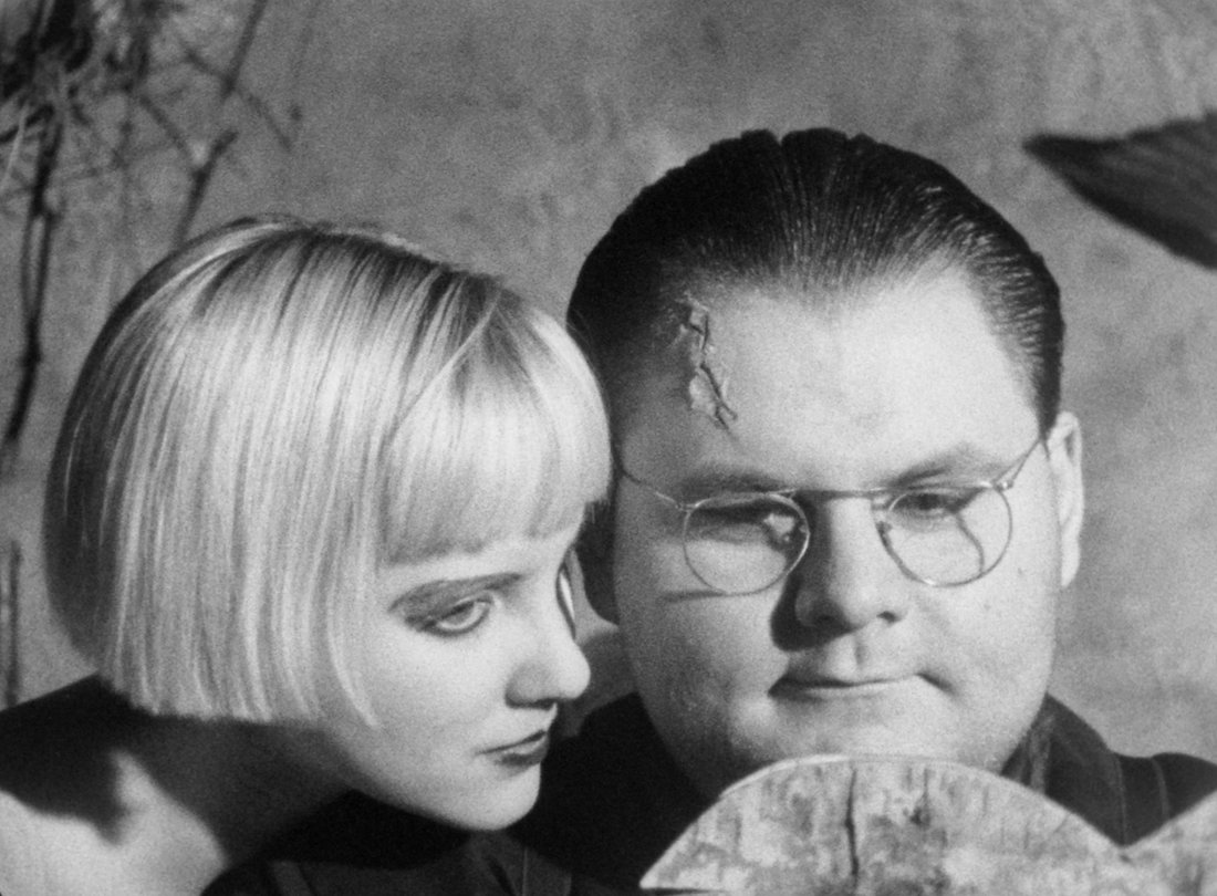 Still from the film Tales from the Gimli Hospital. A young woman and man are seen from the neck up, standing closely together. She is a white woman with a short blonde bob haircut. He has short, slicked back hair. He is wearing wire-rimmed glasses and has a prominent scar on one side of his forehead. They are both staring at something circular but otherwise unidentifiable. 