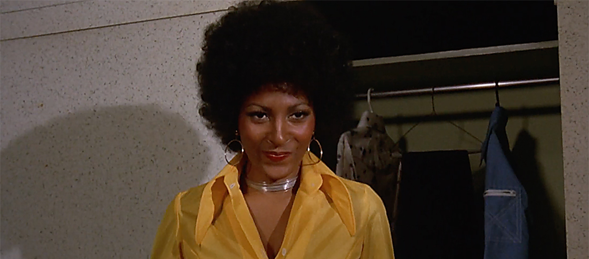 Pam Grier from the chest up in front of a closet. She smirks and wears a yellow button-down shirt. Her hair is styled in an afro.