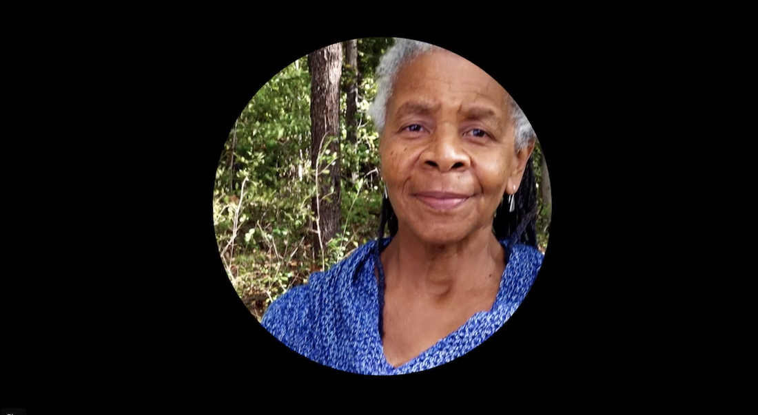 A circular image of dancer-choreographer Bebe Miller surrounded by a a black rectangular frame. She is a Black woman with white and gray dredlocks. She is wearing a blue textured top.  She is standing a little left of center and staring at the camera with a slight smile. She is outside in nature, with trees in the background.