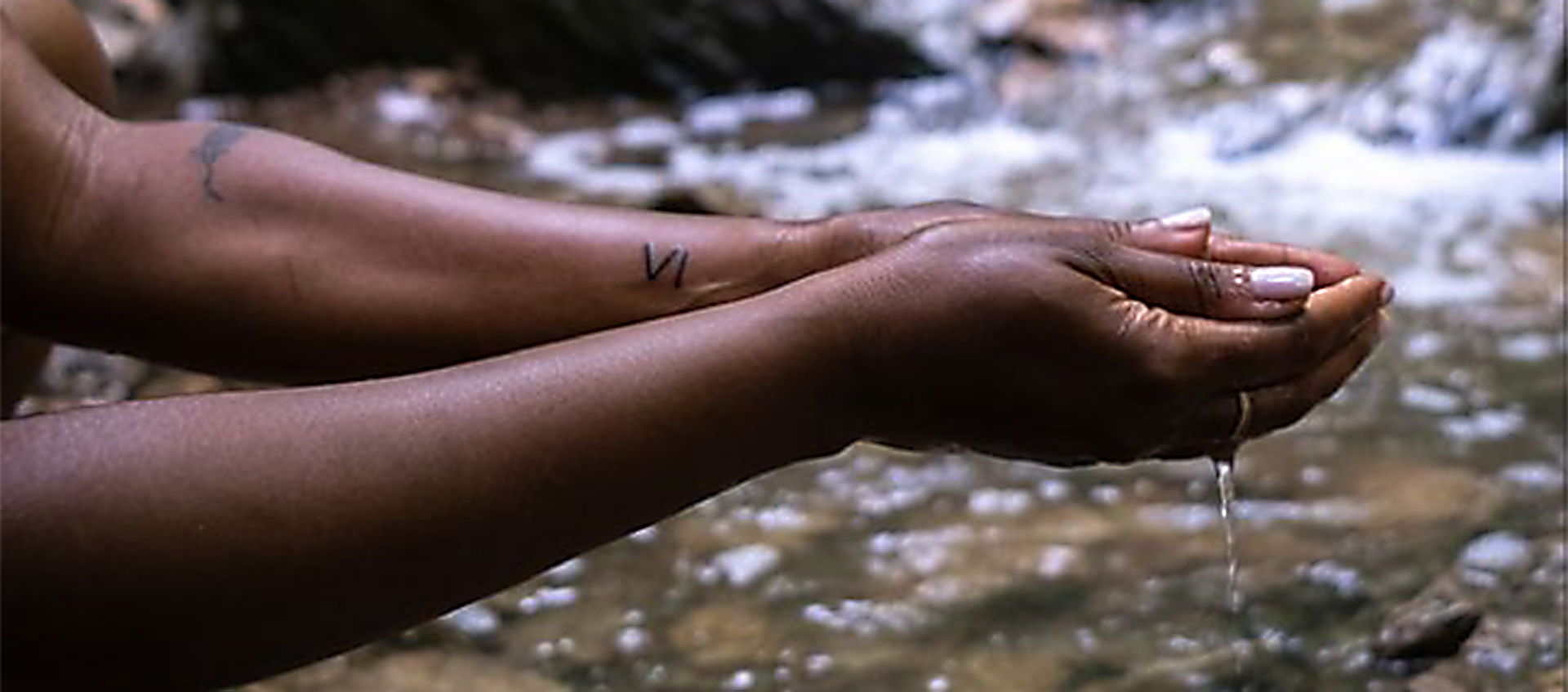 A person’s arms with dark brown skin are outstretched, and their hands are cupping water from a stream visible in the background