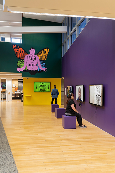 Three monitors on a purple wall, another wall is painted yellow and green with a mural of a person with butterfly wings.
