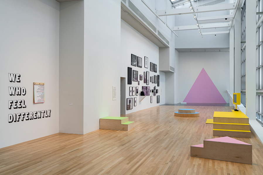 Gallery with large pink triangle in the distance and four geometric platforms painted in pastel colors. Left wall has a group of photographs and text “We Who Feel Differently.”