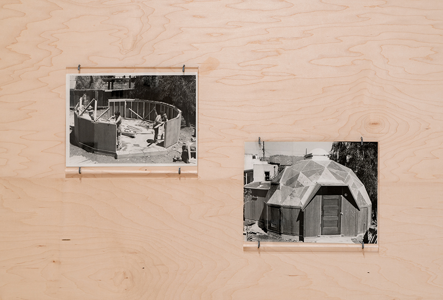 Two black-and-white photographs hung on plywood panels. The photograph at left shows three women working on a building site, where they are framing a circular structure. The photograph at right shows the completed structure, a dome home with a round skylight. 