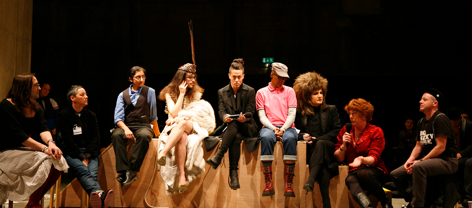 Nine people seated on a geometric wood platform. The second person from the right holds a microphone. 