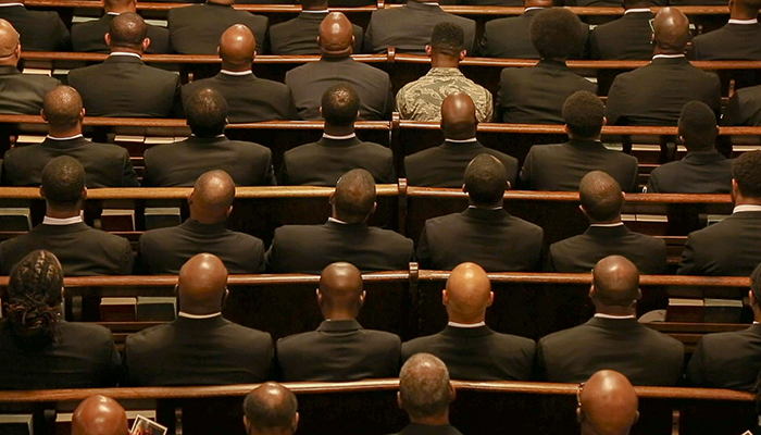 several people in dark suits, one in military fatigues, seated in pews