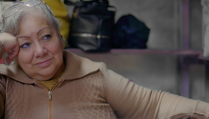 A woman with gray hair sits with her right hand resting on the side of her face. A pair of eyeglasses sit atop her head.