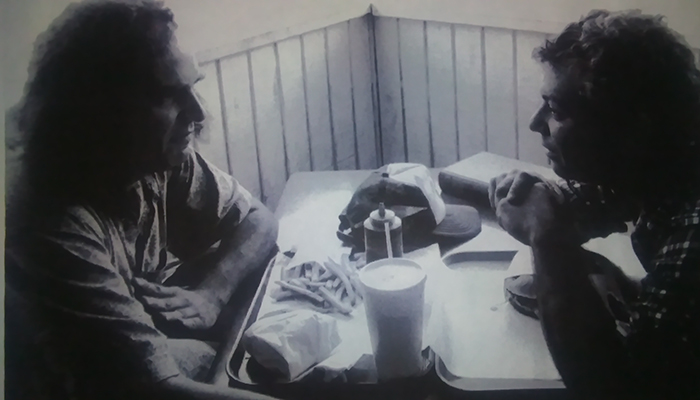 Two men sitting at a table facing each other with a hamburger and fries between them. 