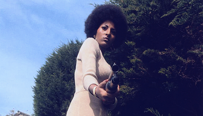 Pam Grier stands in front of trees and holds a shotgun pointed directly at the camera