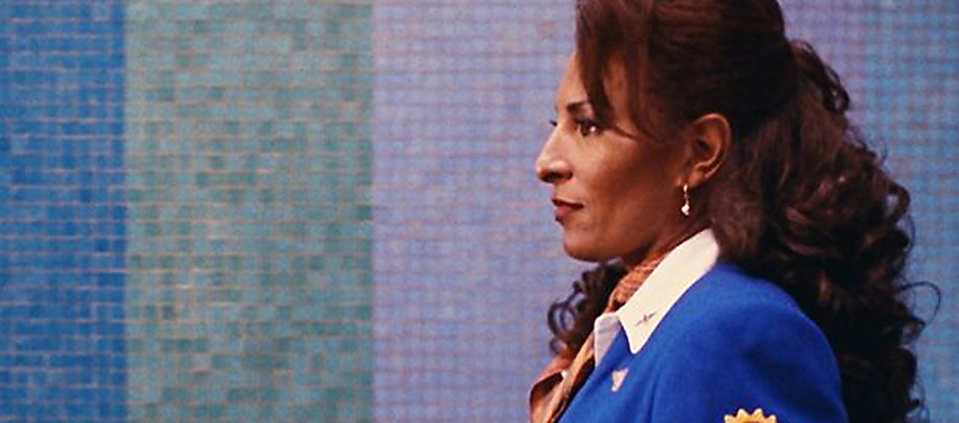 Pam Grier in profile wearing a bright blue flight attendant uniform with white collar. Her hair is long and hangs over her shoulders. She stares straight ahead in front of a blue tiled wall.