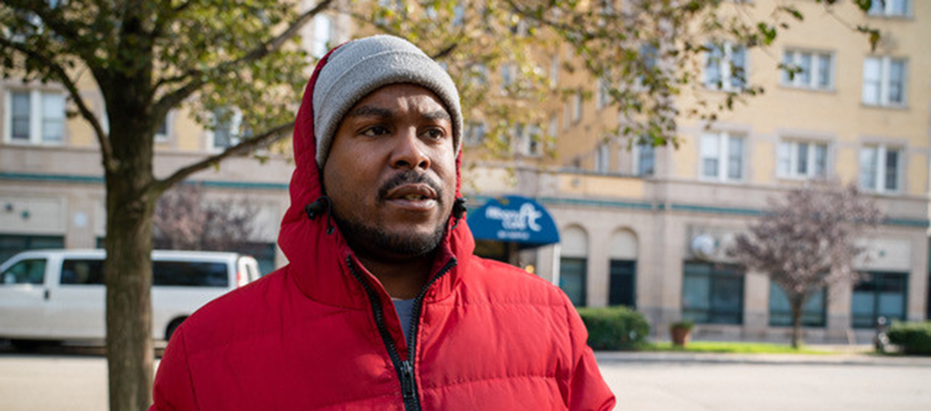 A Black man wearing a red puffy coat and gray knit cap stands in a park.