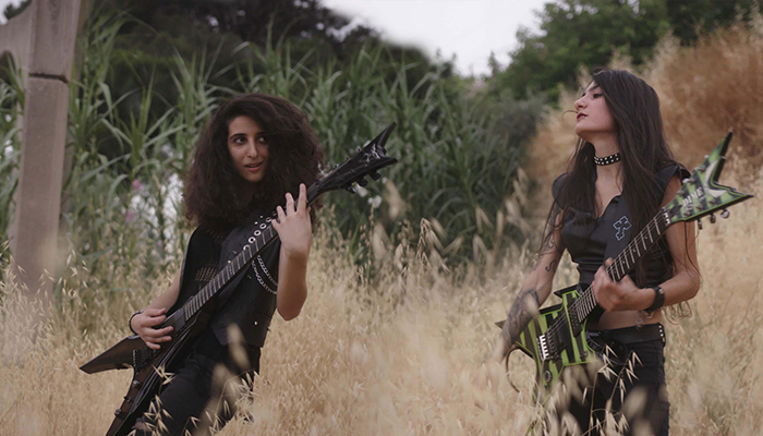 Two girls each holding guitars stand in a field playing music. Both have dark, long hair and wear dark clothes.