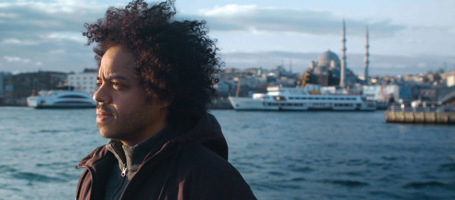 A man in a brown jacket stands in profile in front of a body of water. In the background are several boats. The man has dark, curly hair and a moustache