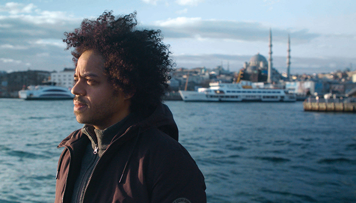 A man in a brown jacket stands in profile in front of a body of water. In the background are several boats. The man has dark, curly hair and a moustache