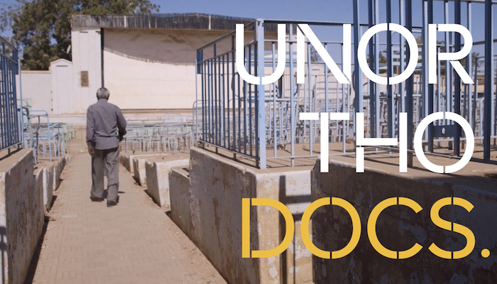 A full-body shot of a Black man with short white hair. He is outdoors, walking away from the camera. There is a small beige building in front of him. He walks a paved path between concrete risers, topped with metal bars. A text graphic spelling out "Unorthodocs" is on the right side of the frame.