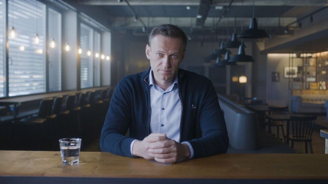 A white, 40-ish man with short brown hair sits at a wooden bar, with an empty restaurant in the background. He wears a light collared shirt and dark blue, open-front zip-up sweater. He's looking directly at the camera with his hands folded in front of him. A shot glass of clear liquid is to his right.