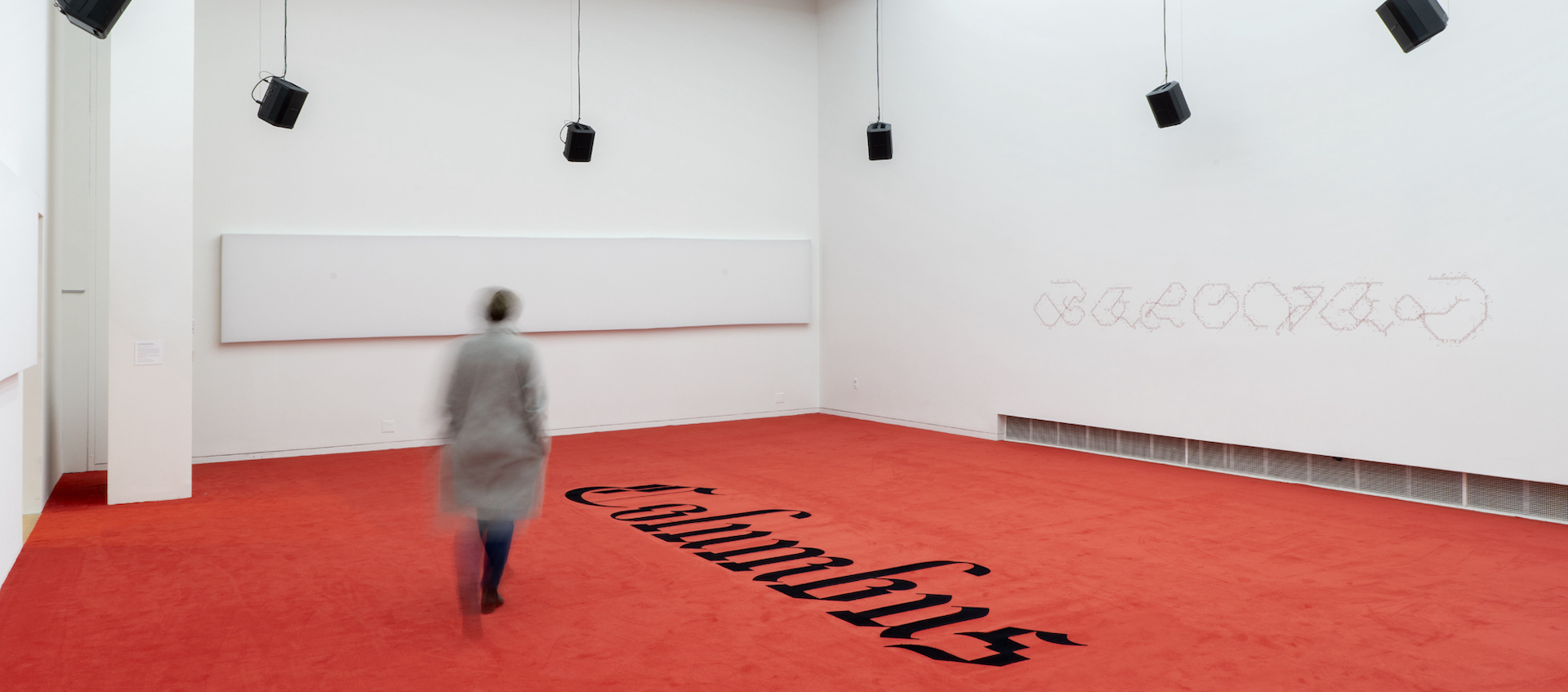 A blurred female figure moves away from the camera through a white-walled gallery space. There are small speakers hanging from the ceiling at points around the room. The floor is covered by a red carpet with the word "Columbus" in old English-style text at the center.