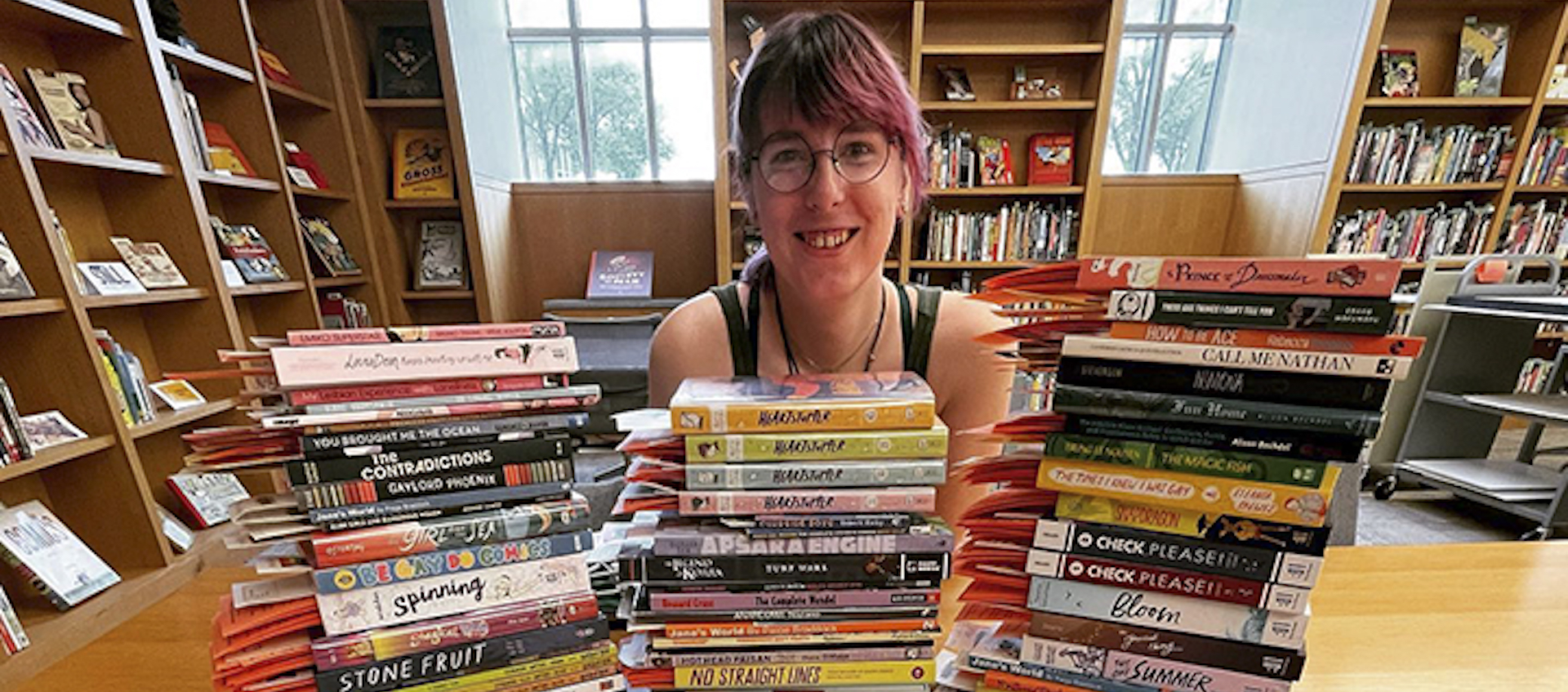 Ohio State research fellow Maggie Dahlstrom is at the center of the image, seen from the shoulders up. They are sitting in a library with book-filled shelves lining the walls and two windows in the background. Maggie has pink hair with bangs and it is pulled back. There are piles of graphic novels about LGBTQ+ youth on a wooden table in front of them.