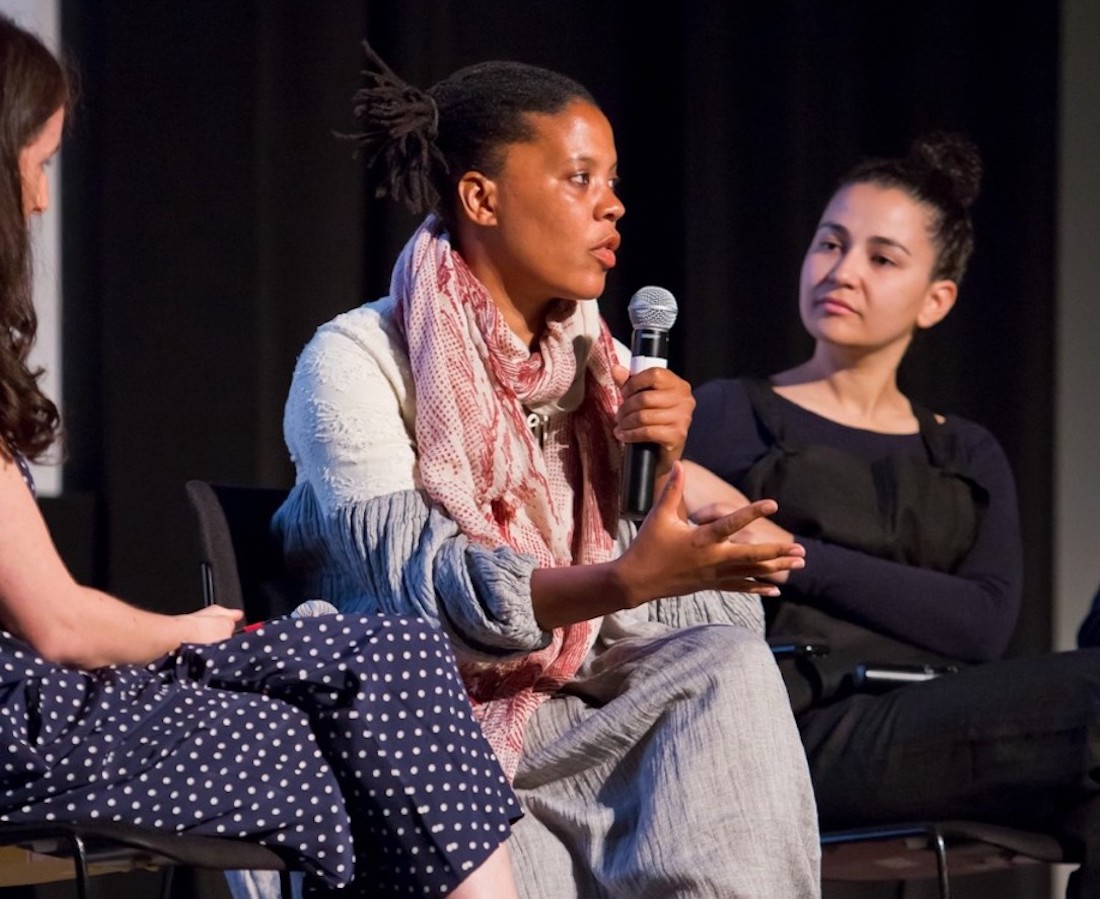  Xaviera Simmons sits as she speaks into a microphone in her hand. She is a Black woman with short dreadlocks pulled back into a ponytail, wearing a loose white and blue dress and a long pink scarf. To the left, artist Carmen Winant is in partial view, a white woman with long dark hair and a blue polka dot dress, also sitting. Artist Bethany Collins, a woman of mixed race wearing a black top and pants, with hair pulled back into a ponytail, is sitting to the right. Both women are looking at Simmons.