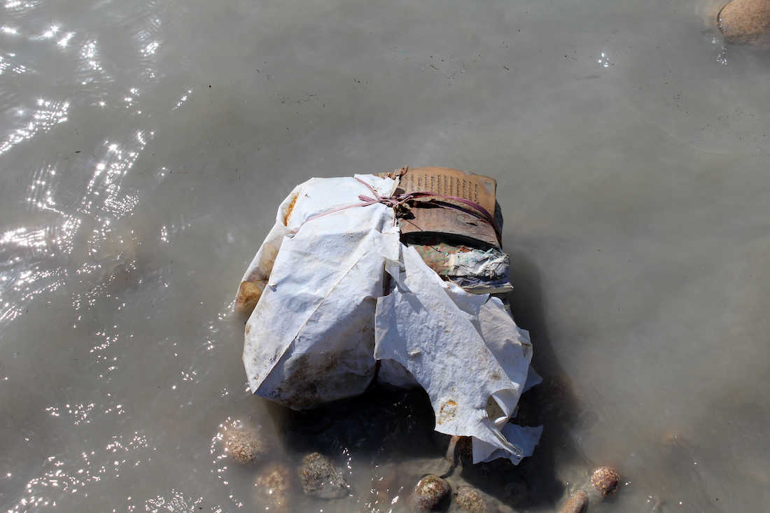 This photograph is an image of a Quran wrapped in leather, cloth, and twine. The Quran is sitting on rocks in a shallow water. It was found floating on the Indus River.