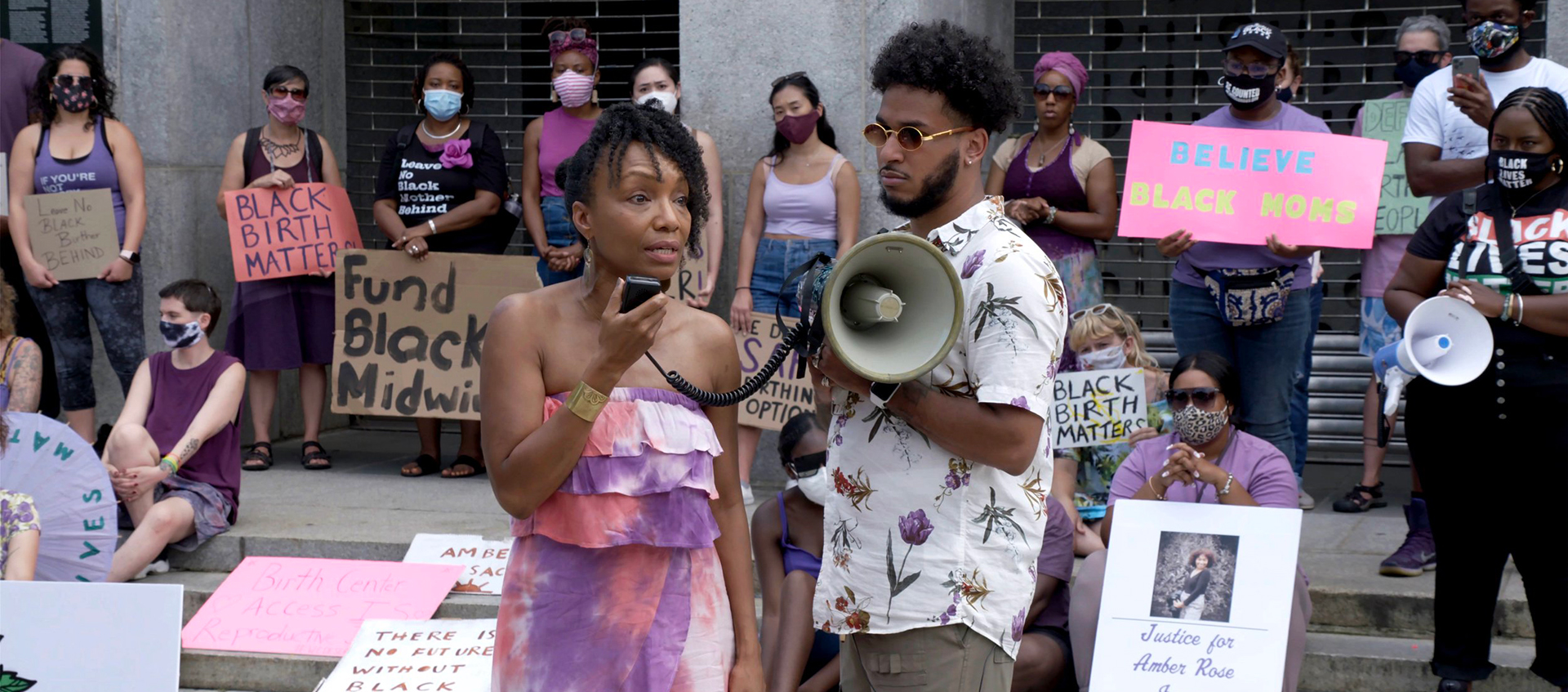 A group of people holding protest signs about justice for Black mothers. Two Black people stand in front of the group holding a megaphone.