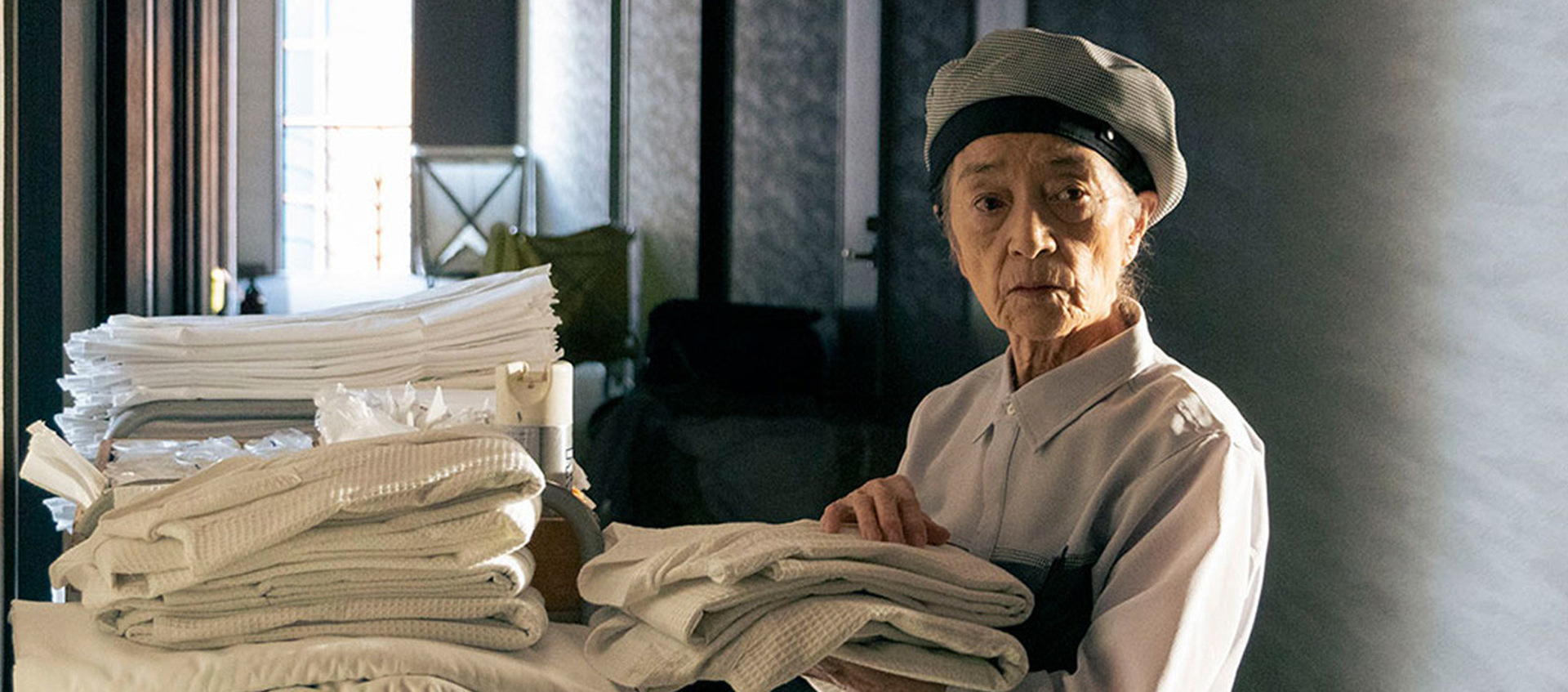 An older Japanese person in a work uniform holds folded laundry and looks into the distance beyond the viewer.