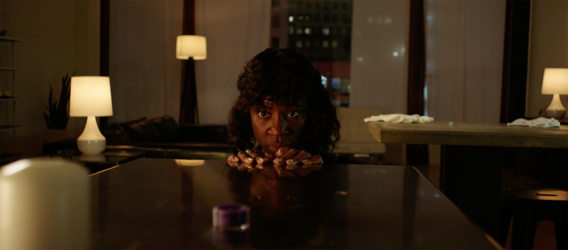 A Black woman peers over a table with her fingertips resting on its surface.  Behind her is a living room lit with lamps.