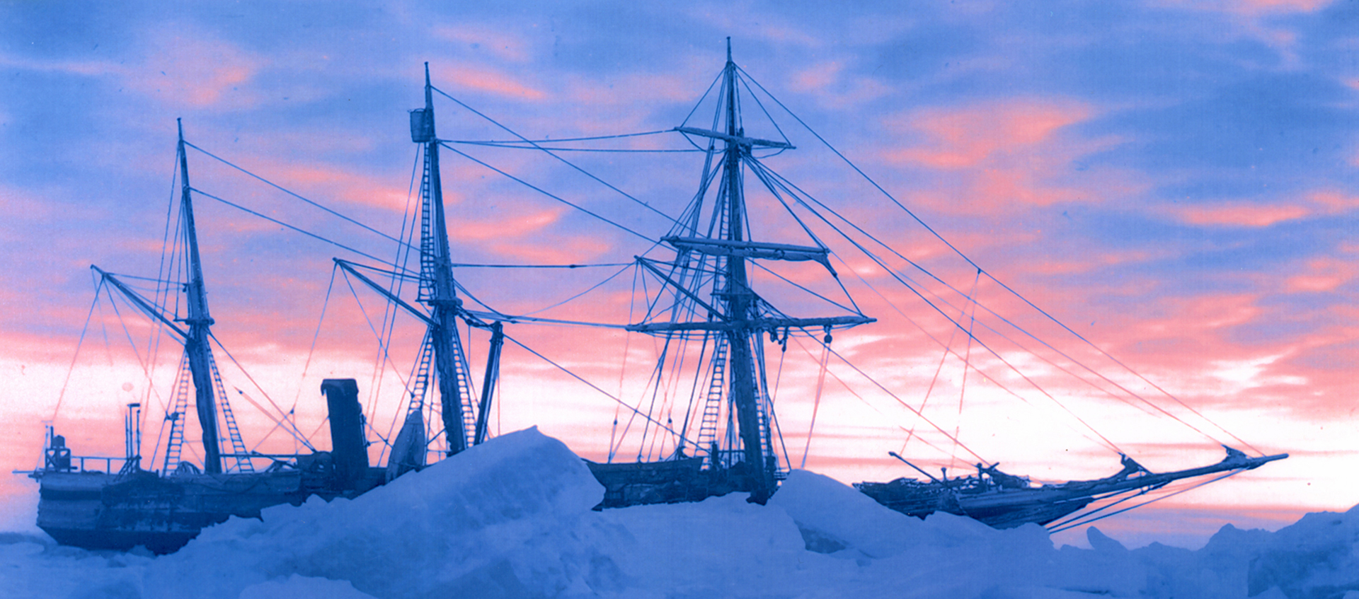 Side view of a sailing vessel, partially obscured by pack ice, against a blue and pink sky.