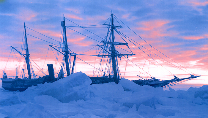Side view of a sailing vessel, partially obscured by pack ice, against a blue and pink sky. 