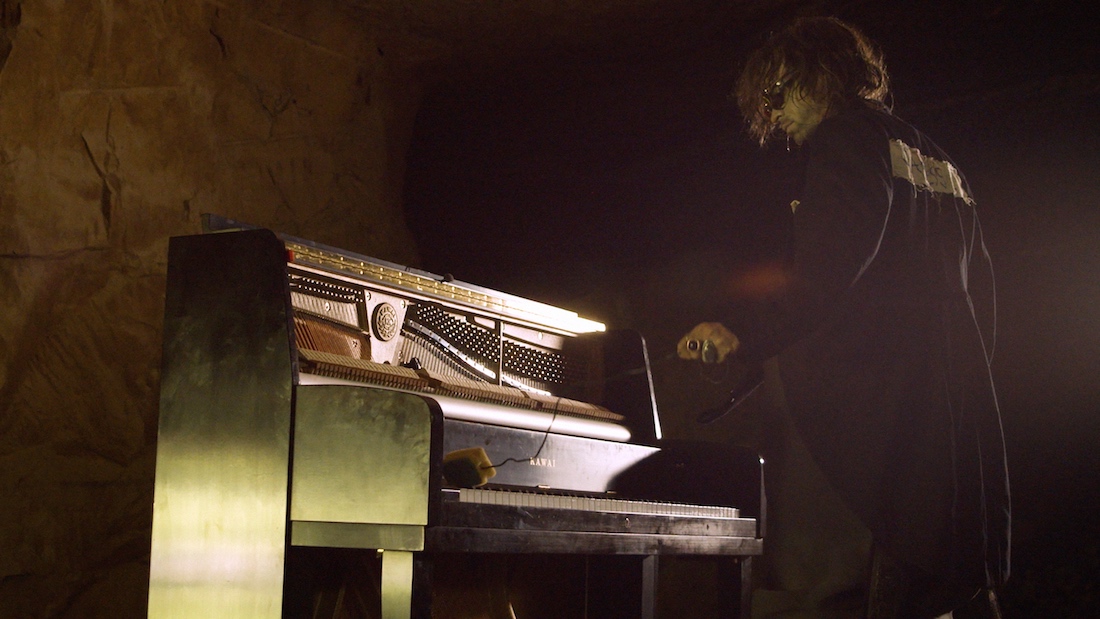 A person in sunglasses and a dark coat faces a piano