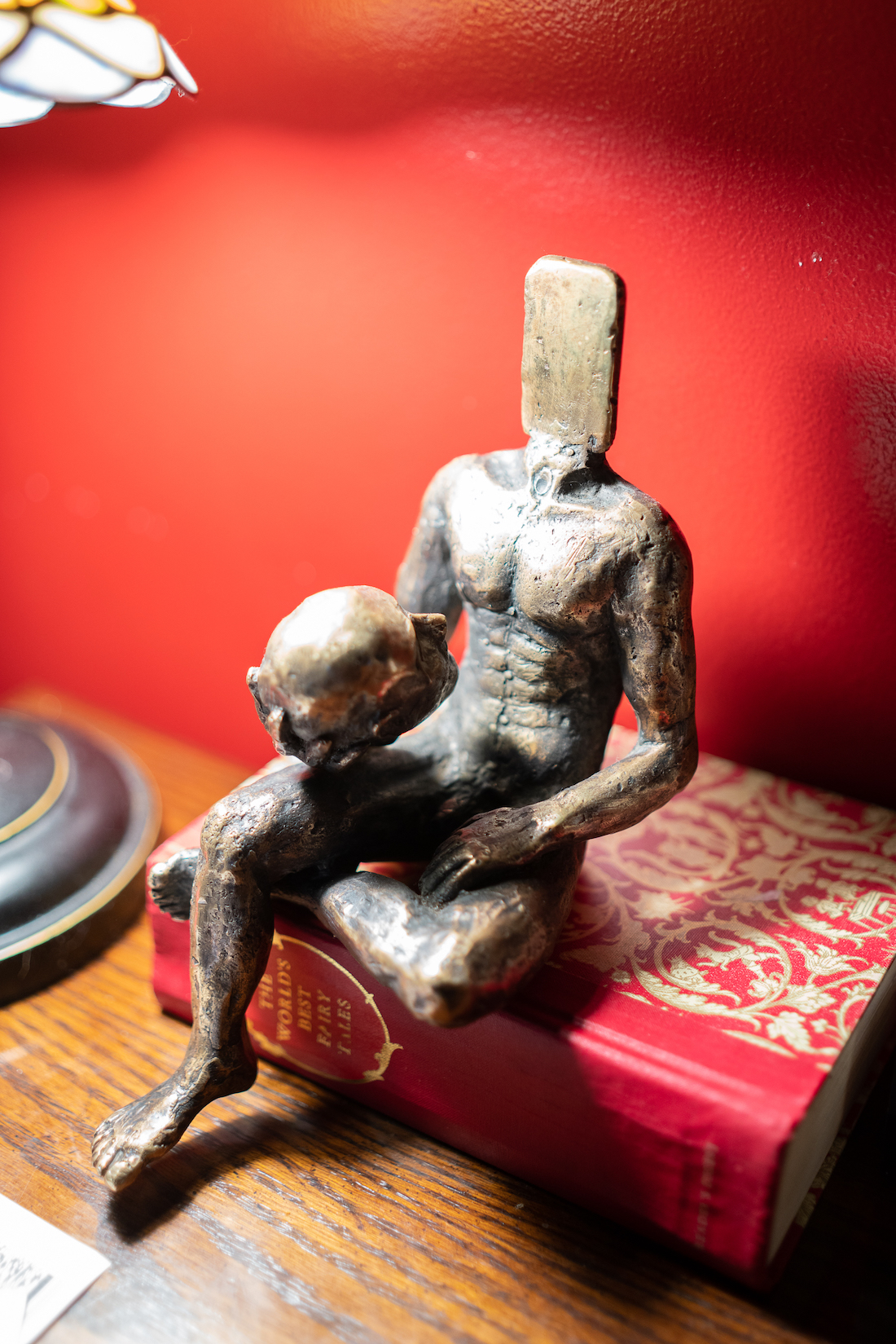 A small bronze figure of a nude man seated. He's holding in one hand his disembodied head. Where his head should be is an oversized smartphone. The piece is placed on a hardcover book with a red cover, which is sitting on a wooden table next to a Tiffany-style lamp.