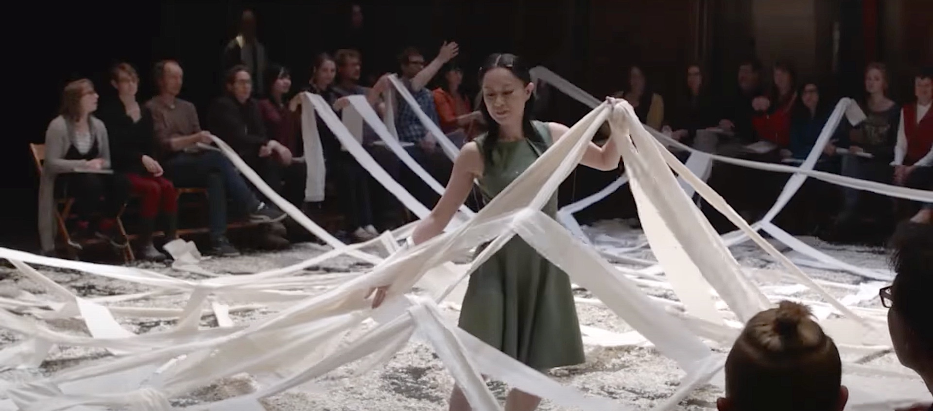 Still from a performance of the theater is a blank page. A woman in a gray sleeveless dress collects narrow strands of paper that crisscross across the performance area. Audience members sit around the performance area and hold the other ends of the paper strands.