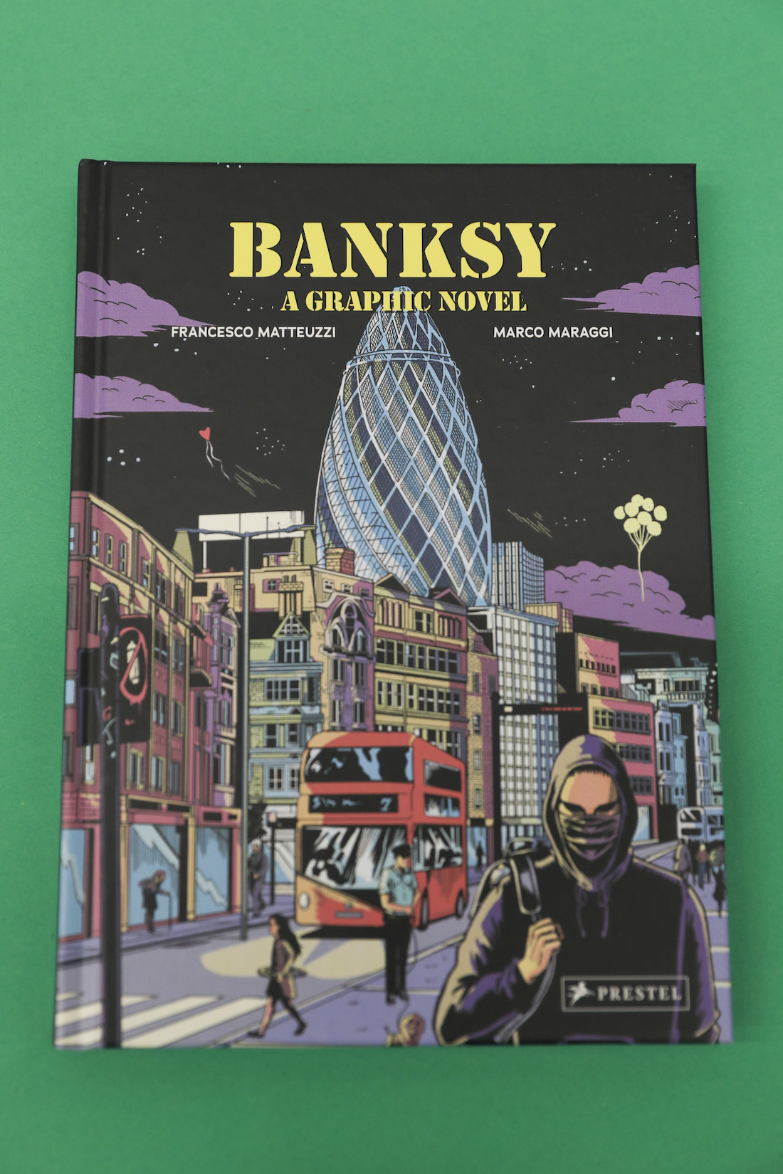 Cover art for Banksy: A Graphic Novel. A male figure in all black with a hoodie and a face mask looks straight ahead in the foreground. A cityscape under a night sky with purple clouds is in the background.