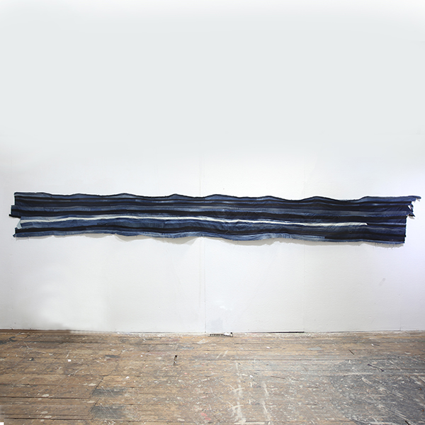 Eye-level shot of a long horizontal strip of blue, black, and white denim fabric sewn together, hanging on a white gallery wall.