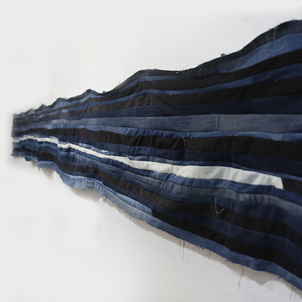 Side-angle shot of a long horizontal strip of blue, black, and white denim fabric sewn together, hanging on a white gallery wall.