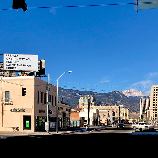 Wide shot of a white billboard atop a commercial building with black text that reads, "I REALLY LIKE THE WAY YOU RESPECT NATIVE AMERICAN RIGHTS."