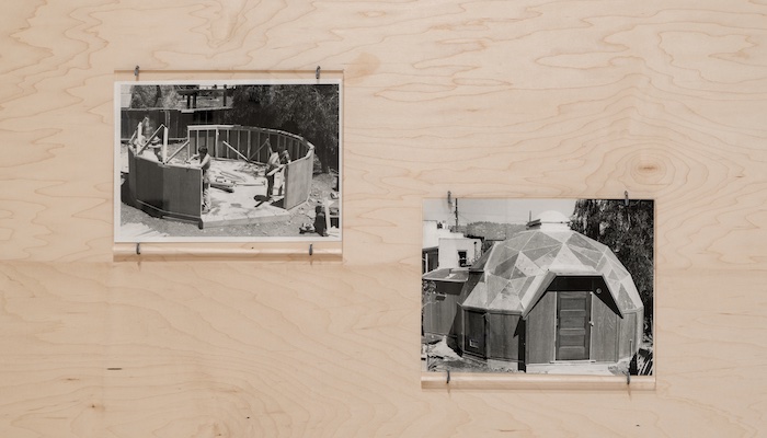Two black-and-white photographs hung on plywood panels. The photograph at left shows three women working on a building site, where they are framing a circular structure. The photograph at right shows the completed structure, a dome home with a round skylight. 