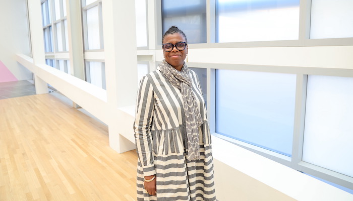 Gaëtane Verna stands smiling in a bright gallery space lined with film-covered windows and white metal grid work. She is a Black woman with pulled back braided hair. She wears black-rimmed glasses, a gray and white striped dress and a gray and white scarf.